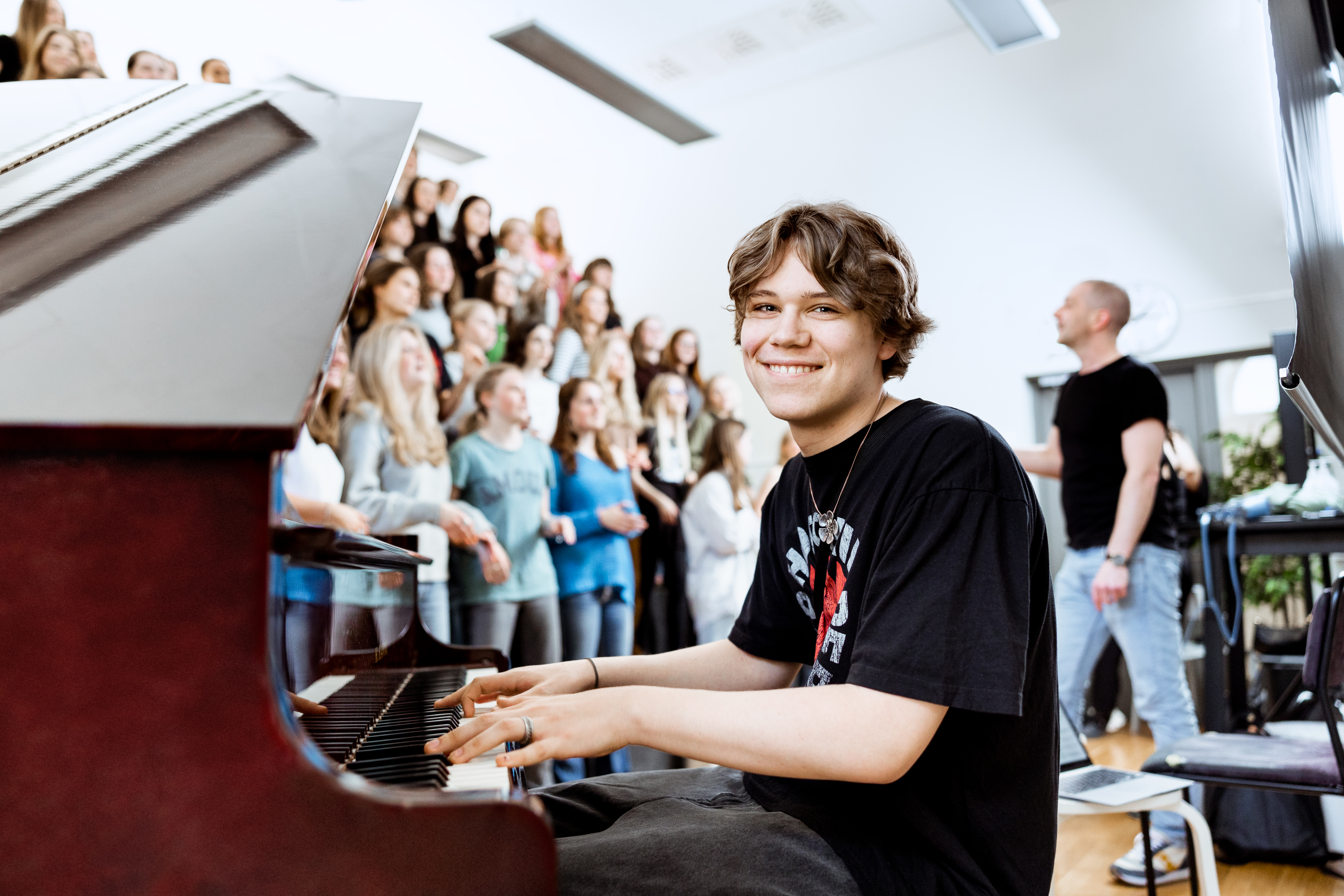 Elev tittar leende in i kameran samtidigt som han spelar piano. I bakgrunden syns en kör.